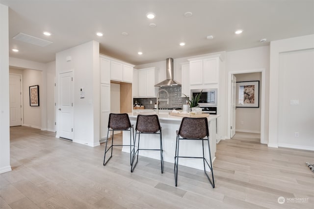 kitchen featuring wall chimney range hood, appliances with stainless steel finishes, a kitchen island with sink, white cabinets, and light wood-type flooring