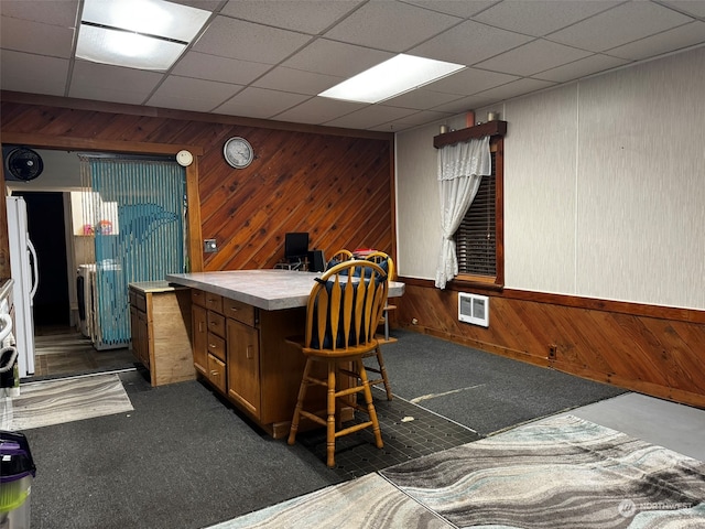 interior space featuring a drop ceiling, dark colored carpet, wooden walls, and white fridge