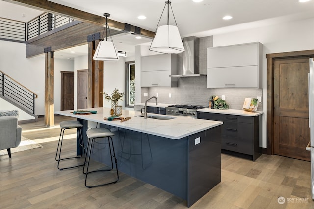 kitchen with sink, wall chimney range hood, pendant lighting, light hardwood / wood-style floors, and a kitchen island with sink