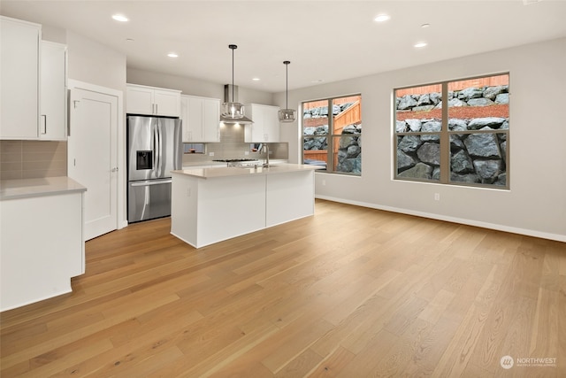 kitchen with stainless steel fridge, hanging light fixtures, wall chimney exhaust hood, white cabinetry, and a kitchen island with sink