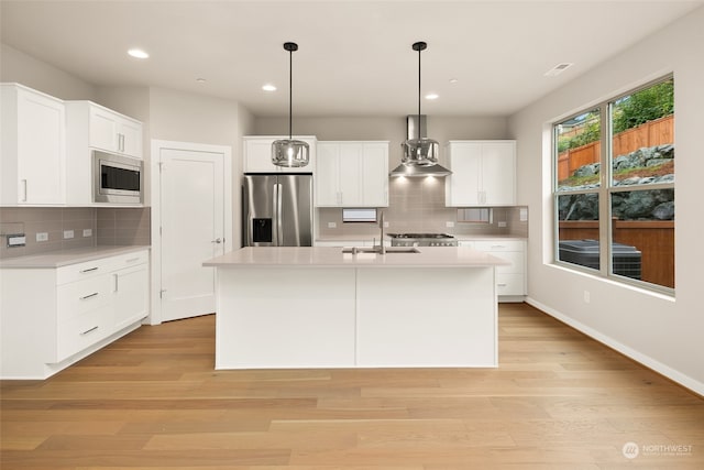 kitchen with pendant lighting, white cabinets, appliances with stainless steel finishes, wall chimney exhaust hood, and a center island with sink