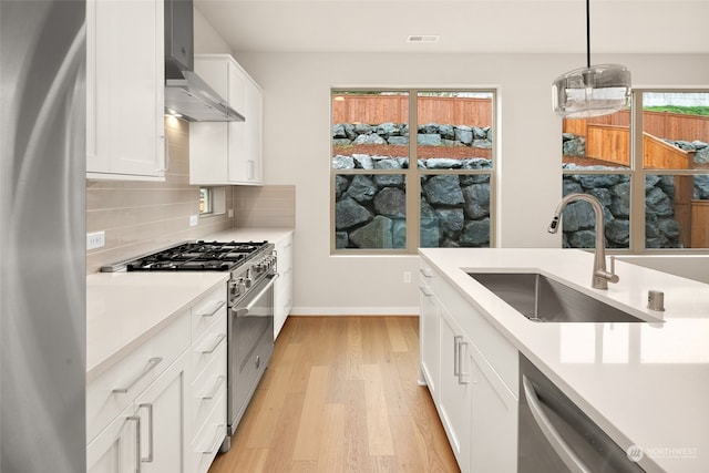 kitchen with appliances with stainless steel finishes, decorative light fixtures, wall chimney exhaust hood, sink, and white cabinetry