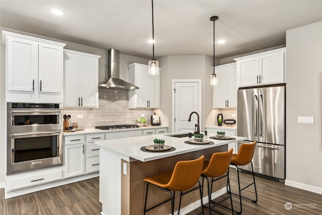 kitchen featuring stainless steel appliances, a kitchen island with sink, white cabinets, wall chimney range hood, and pendant lighting