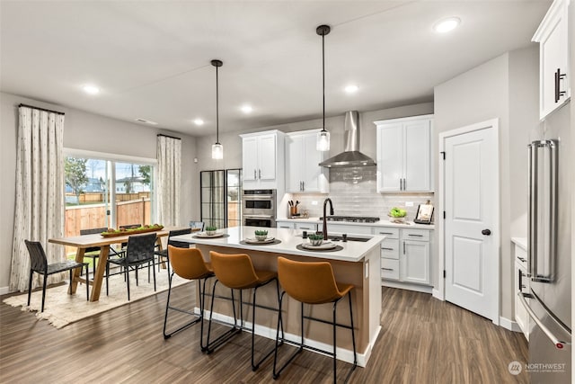 kitchen featuring wall chimney range hood, white cabinetry, a kitchen island with sink, and appliances with stainless steel finishes