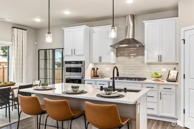 kitchen with stainless steel appliances, wall chimney exhaust hood, a kitchen island with sink, white cabinets, and pendant lighting