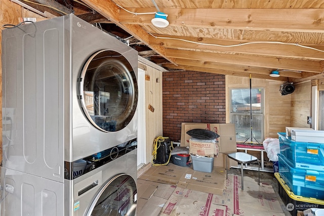 laundry area featuring wooden walls, stacked washer and clothes dryer, and brick wall