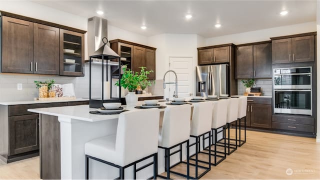 kitchen featuring decorative backsplash, appliances with stainless steel finishes, light wood-type flooring, a breakfast bar, and an island with sink