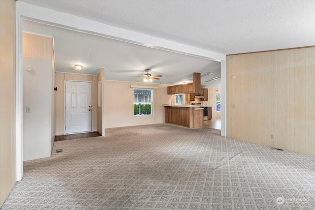 unfurnished living room with ceiling fan, wooden walls, beamed ceiling, a textured ceiling, and light carpet