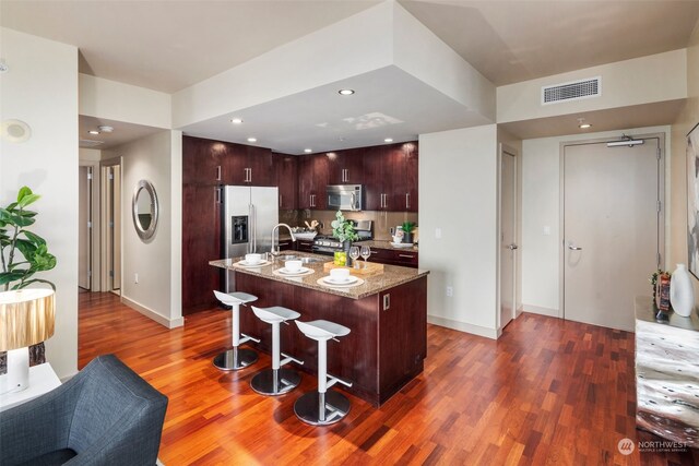 kitchen with stainless steel appliances, a center island with sink, a breakfast bar, light stone countertops, and dark wood-type flooring