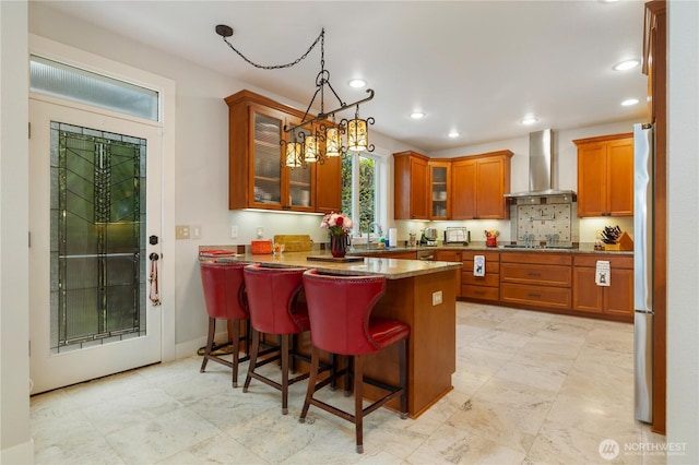 kitchen with a breakfast bar, a sink, wall chimney range hood, brown cabinetry, and glass insert cabinets