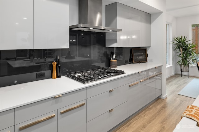 kitchen featuring wall chimney range hood, gray cabinets, light wood-type flooring, tasteful backsplash, and stainless steel gas cooktop