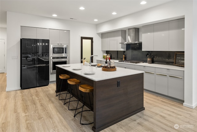 kitchen with appliances with stainless steel finishes, a kitchen breakfast bar, wall chimney exhaust hood, sink, and an island with sink