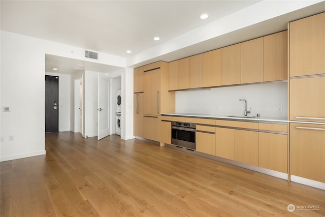 kitchen featuring sink, light brown cabinets, light hardwood / wood-style flooring, stacked washer / dryer, and oven