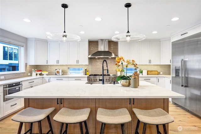 kitchen featuring stainless steel appliances, wall chimney range hood, an island with sink, decorative light fixtures, and white cabinets