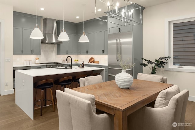 kitchen with wall chimney range hood, sink, hanging light fixtures, and an island with sink