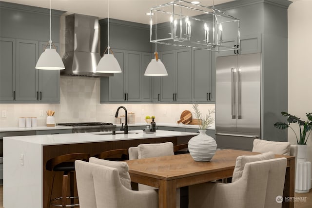 kitchen featuring pendant lighting, a center island with sink, wall chimney range hood, built in refrigerator, and decorative backsplash