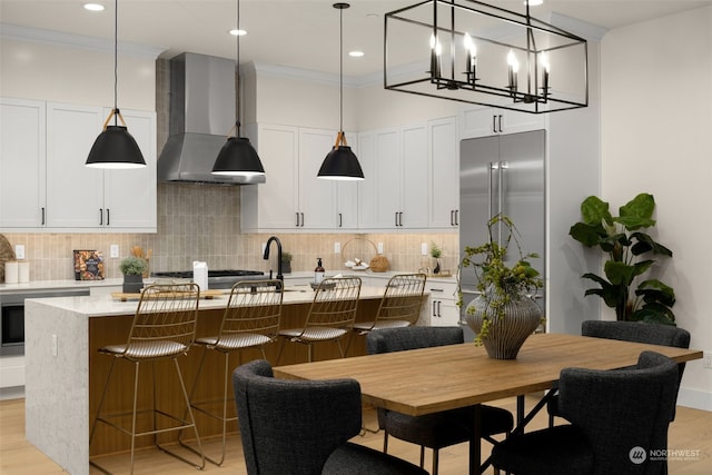 kitchen with white cabinets, wall chimney exhaust hood, an island with sink, and decorative light fixtures