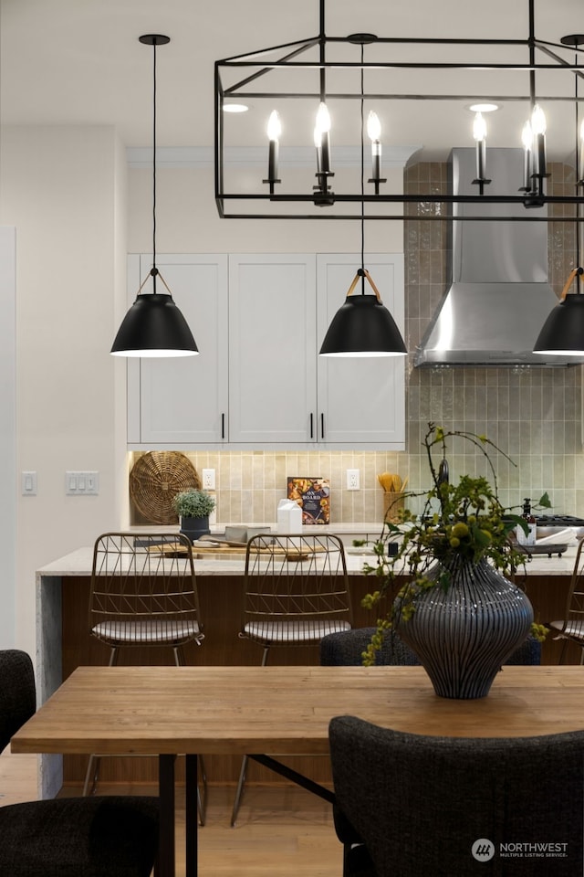 kitchen with wall chimney range hood, backsplash, hardwood / wood-style floors, decorative light fixtures, and white cabinets