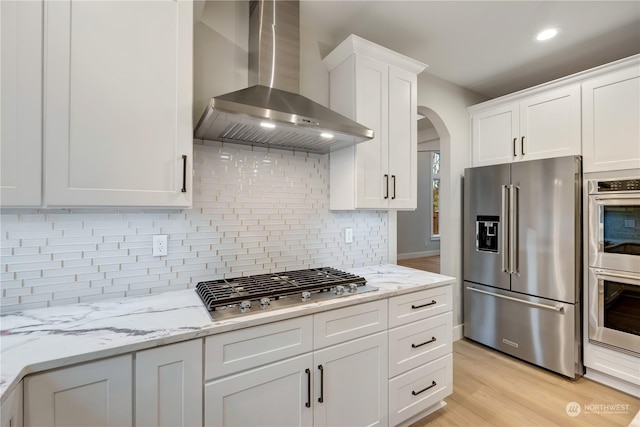 kitchen with white cabinets, wall chimney range hood, light hardwood / wood-style flooring, appliances with stainless steel finishes, and tasteful backsplash