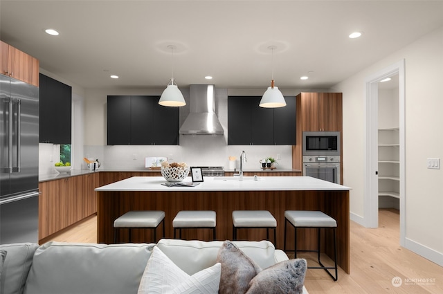 kitchen with pendant lighting, wall chimney range hood, an island with sink, and light hardwood / wood-style flooring