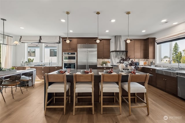 kitchen featuring dark brown cabinets, stainless steel appliances, wall chimney range hood, decorative light fixtures, and a center island