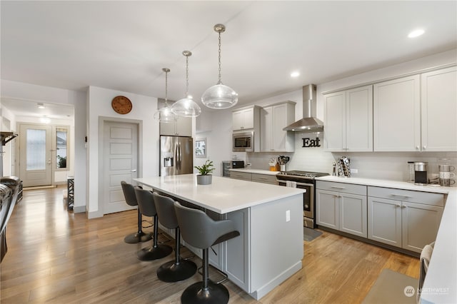 kitchen with a breakfast bar, wall chimney range hood, appliances with stainless steel finishes, decorative light fixtures, and light hardwood / wood-style floors