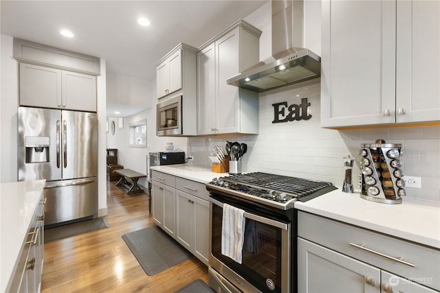 kitchen featuring wall chimney range hood, backsplash, light hardwood / wood-style floors, gray cabinets, and appliances with stainless steel finishes