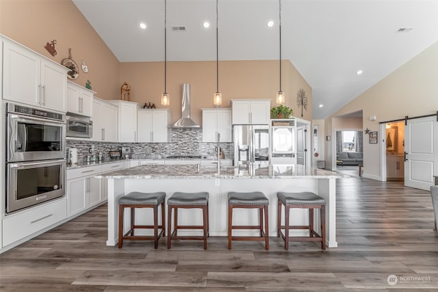 kitchen featuring a kitchen island with sink, high vaulted ceiling, wall chimney exhaust hood, a barn door, and stainless steel appliances