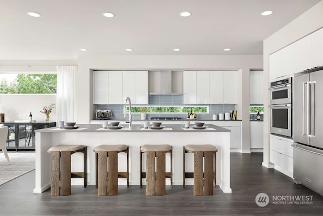 kitchen with wall chimney exhaust hood, stainless steel appliances, a breakfast bar area, and an island with sink