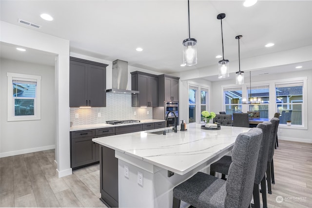 kitchen featuring wall chimney exhaust hood, light stone counters, decorative light fixtures, a center island with sink, and light wood-type flooring