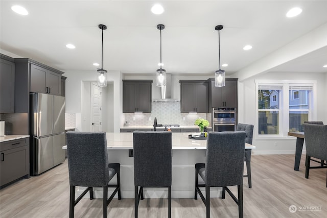 kitchen with pendant lighting, light wood-type flooring, stainless steel appliances, and wall chimney exhaust hood