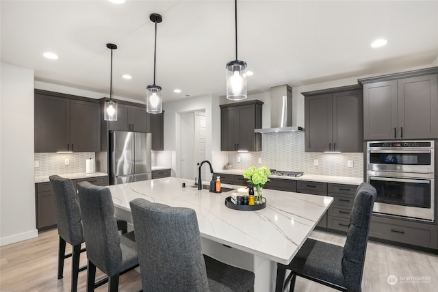 kitchen featuring stainless steel appliances, wall chimney range hood, light hardwood / wood-style flooring, an island with sink, and decorative light fixtures