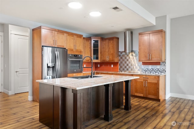 kitchen with dark hardwood / wood-style flooring, wall chimney range hood, a kitchen island with sink, and appliances with stainless steel finishes