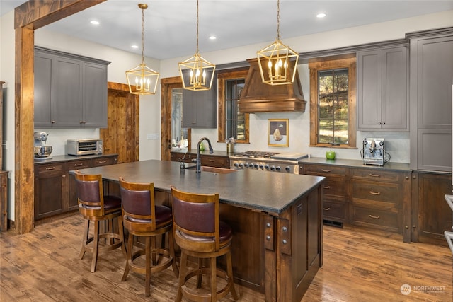 kitchen with sink, light hardwood / wood-style flooring, premium range hood, decorative light fixtures, and a center island with sink