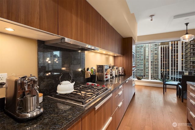 kitchen with stainless steel gas stovetop, dark stone counters, hanging light fixtures, range hood, and light hardwood / wood-style floors