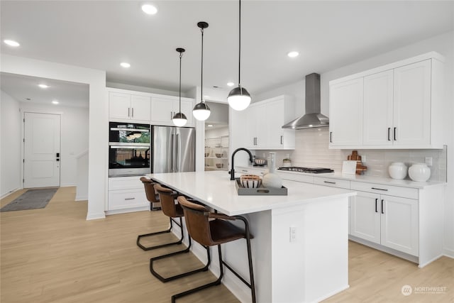 kitchen featuring wall chimney range hood, an island with sink, light hardwood / wood-style floors, white cabinetry, and stainless steel appliances