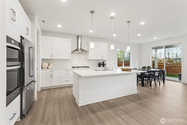 kitchen featuring light wood-style flooring, wall chimney range hood, appliances with stainless steel finishes, light countertops, and decorative backsplash