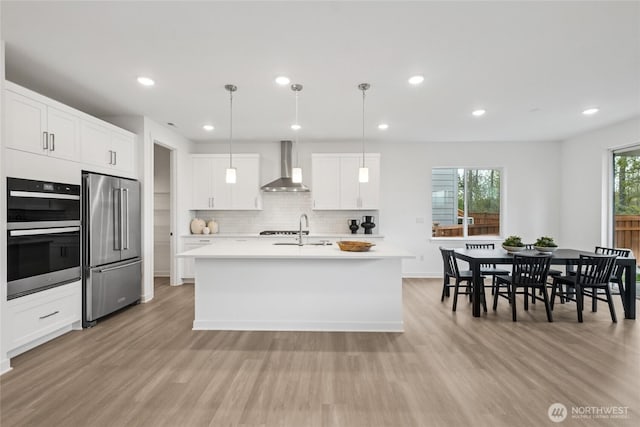 kitchen with stainless steel appliances, light wood finished floors, a sink, and wall chimney range hood