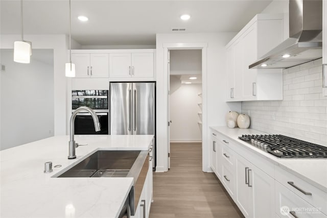 kitchen with wall chimney range hood, white cabinetry, light wood-style floors, stainless steel appliances, and a sink
