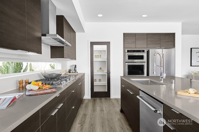 kitchen featuring dark brown cabinetry, wall chimney exhaust hood, light wood-type flooring, and appliances with stainless steel finishes