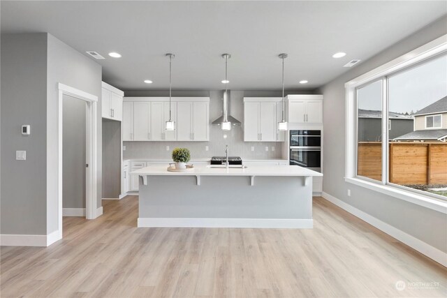 kitchen with an island with sink, pendant lighting, wall chimney range hood, and white cabinets