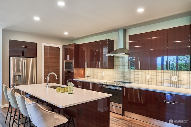 kitchen featuring light wood-type flooring, stainless steel appliances, a kitchen island with sink, and wall chimney exhaust hood