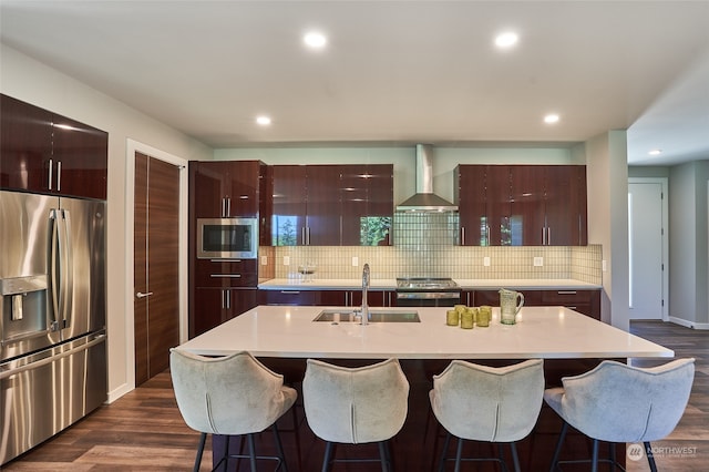 kitchen with sink, wall chimney range hood, dark hardwood / wood-style floors, a kitchen island with sink, and appliances with stainless steel finishes