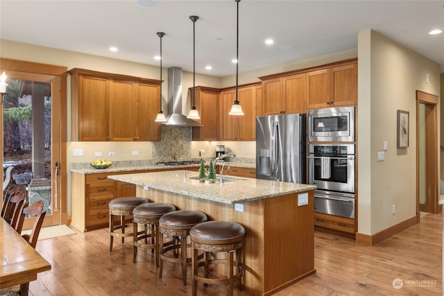 kitchen with a warming drawer, stainless steel appliances, hanging light fixtures, an island with sink, and wall chimney exhaust hood