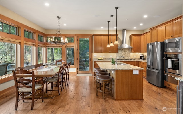 kitchen with pendant lighting, wall chimney exhaust hood, stainless steel appliances, and a kitchen island with sink