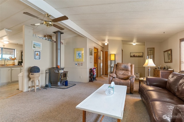 carpeted living room featuring beamed ceiling, a wood stove, ceiling fan, and sink