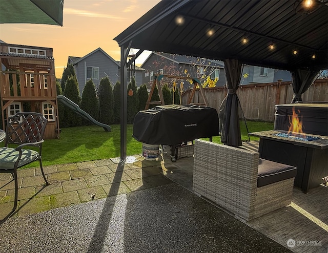 patio terrace at dusk featuring a playground, a grill, a lawn, and a fire pit