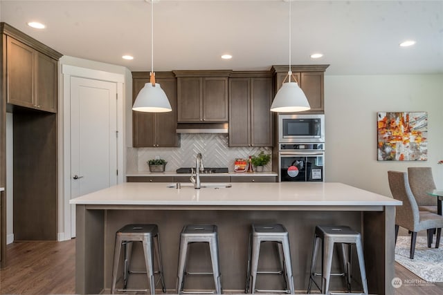 kitchen with appliances with stainless steel finishes, backsplash, pendant lighting, dark hardwood / wood-style floors, and an island with sink