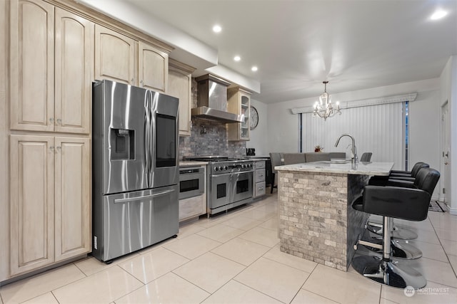 kitchen featuring stainless steel appliances, wall chimney range hood, a chandelier, decorative backsplash, and a center island with sink