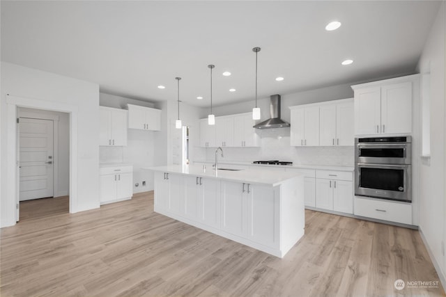 kitchen featuring stainless steel double oven, wall chimney range hood, light hardwood / wood-style flooring, an island with sink, and white cabinets